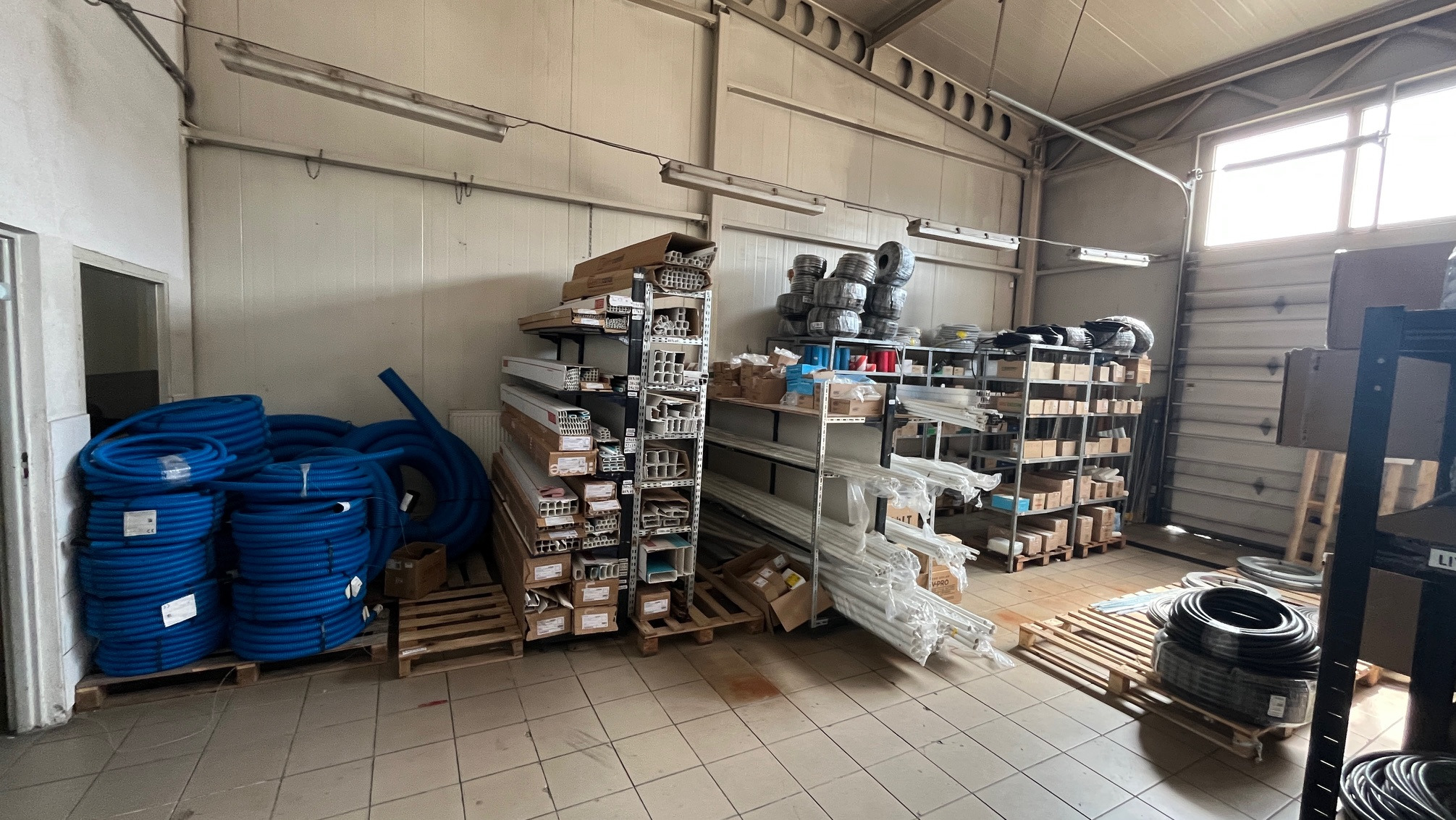 Wnętrze magazynu z rurami instalacyjnymi, profilami i elementami montażowymi na regałach i paletach. Widok na dobrze oświetlone pomieszczenie z różnorodnym asortymentem.