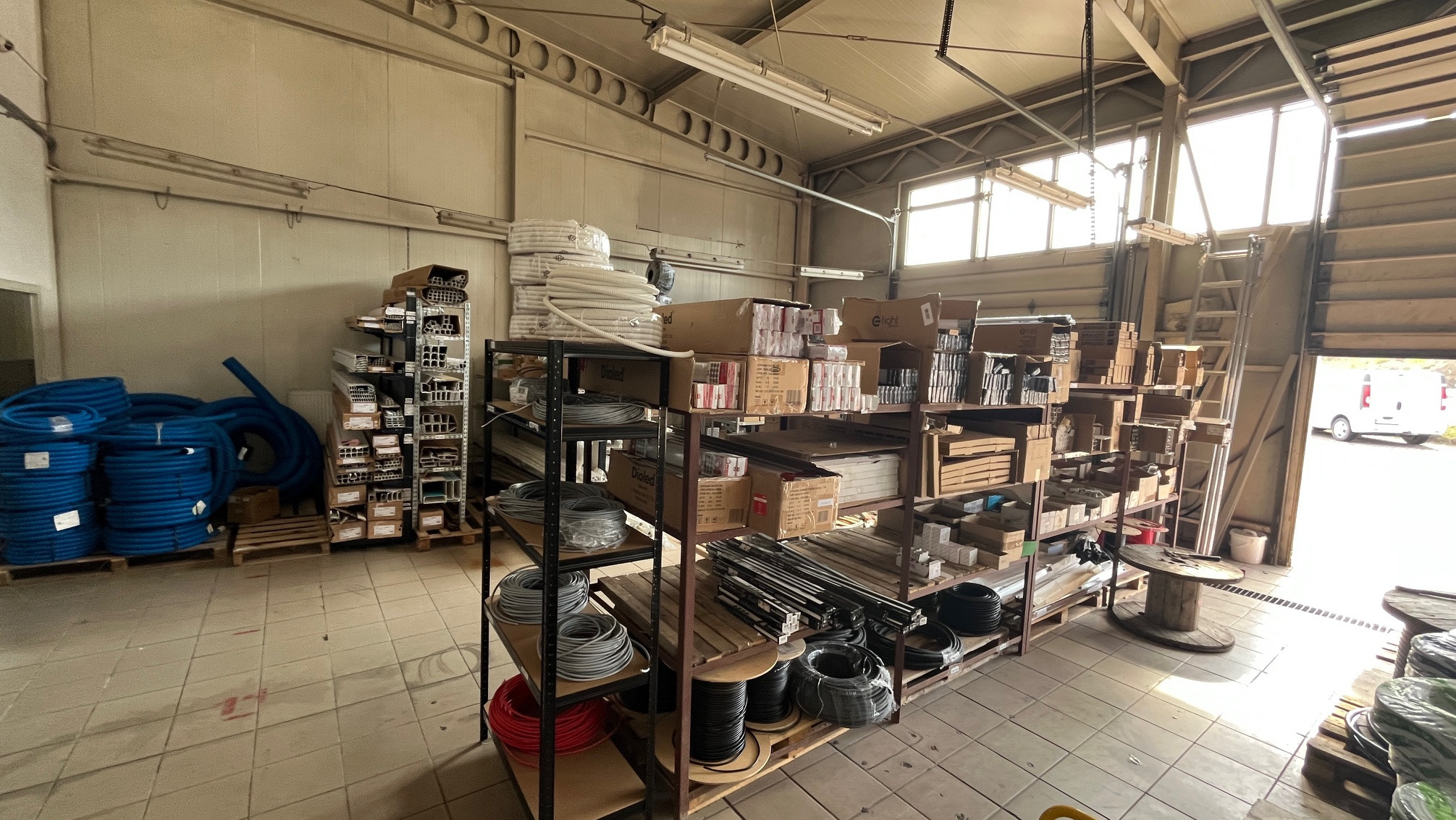 Wnętrze magazynu z rzędami regałów wypełnionych rurami, kablami i akcesoriami instalacyjnymi. Widoczne rolki kabli, kartony i elementy metalowe. Otwarta brama w tle.
