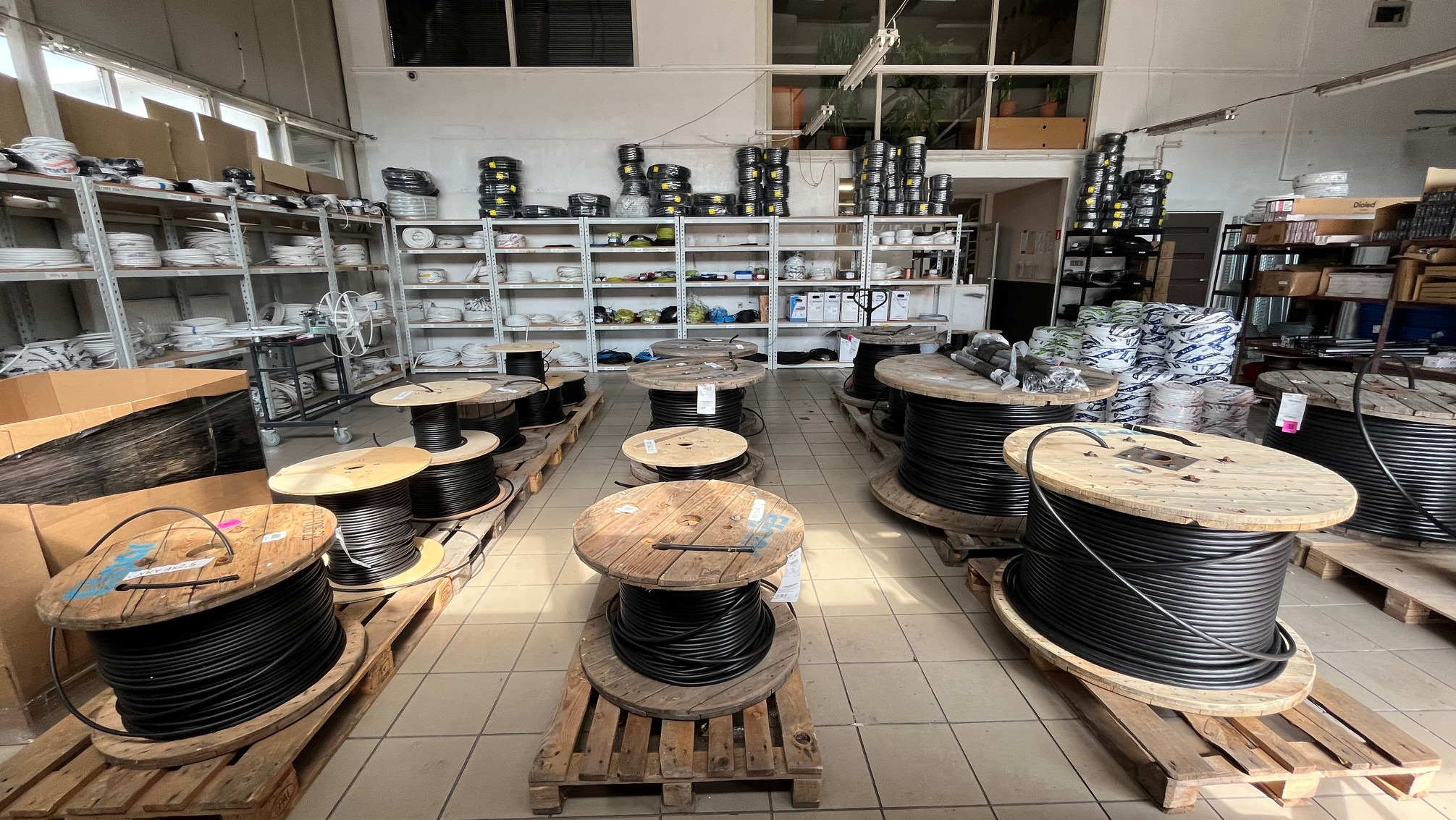 Wnętrze magazynu z kablami na drewnianych szpulach. Rzędy szpul z czarnymi kablami, metalowe regały z różnymi materiałami instalacyjnymi w tle. Widok z perspektywy podłogi.