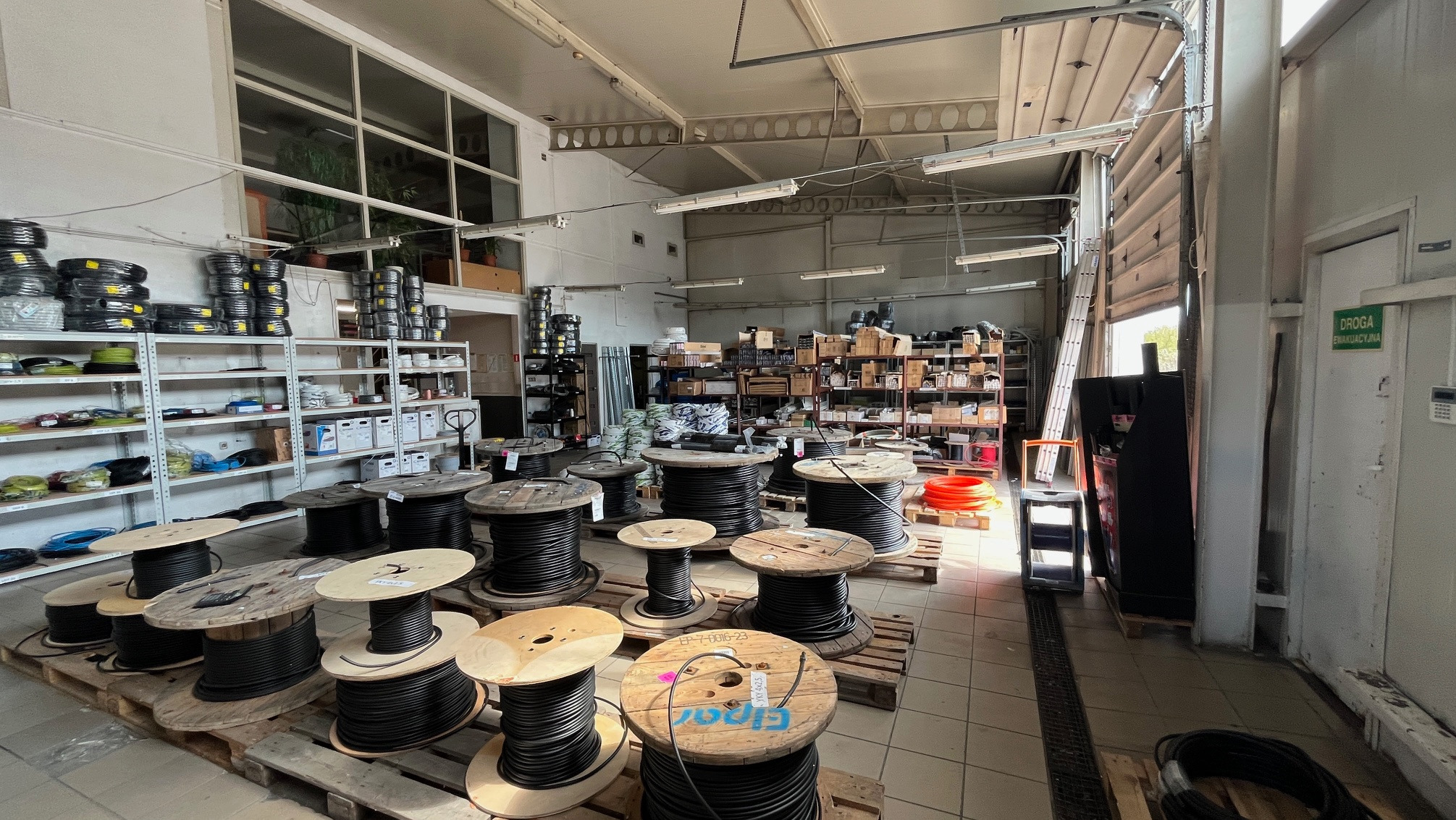 Magazyn kabli: Szpule z czarnym kablem na paletach, regały z kablami w zwojach. Widoczny fragment drzwi ewakuacyjnych i drabina. Oświetlenie magazynowe.