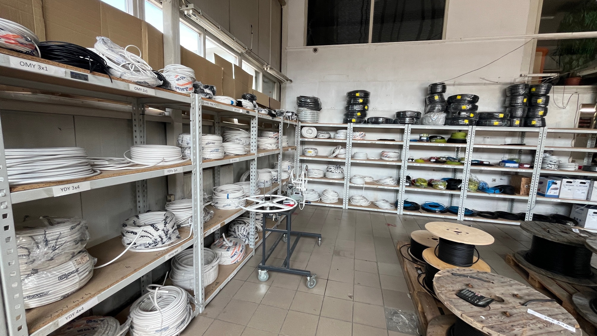 Wnętrze sklepu hydraulicznego: półki z kablami OMY i YDY o różnych przekrojach, szpule z kablami na podłodze, wózek do odwijania kabli. Widok na magazyn z asortymentem.