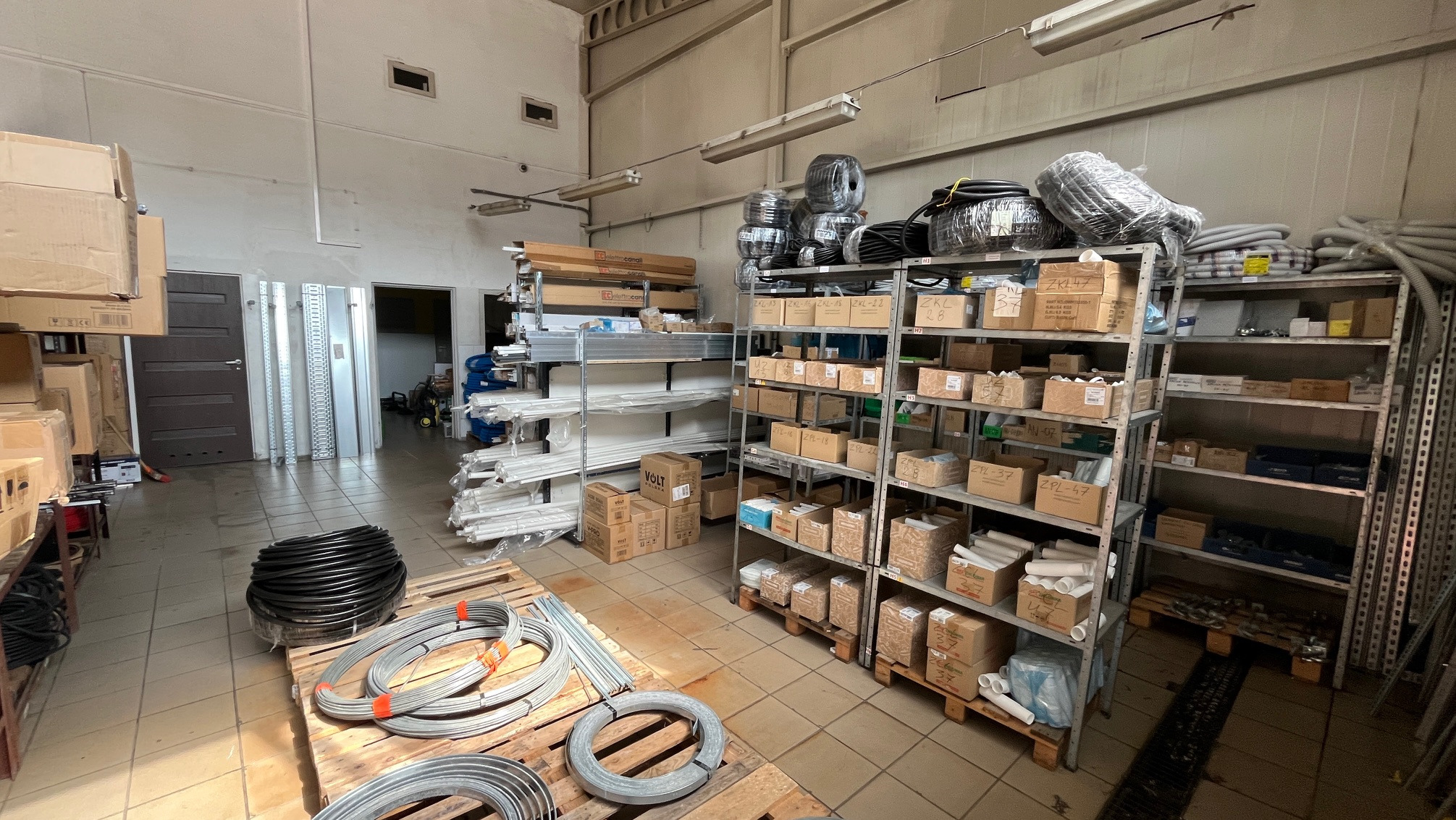 Wnętrze magazynu hydraulicznego z rurami, kształtkami i przewodami na regałach i paletach. Widoczne kartony z oznaczeniami producentów i specyfikacjami.