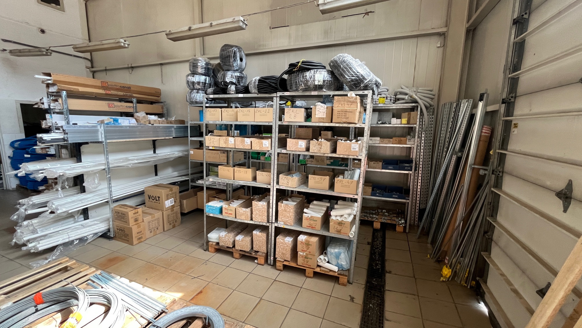 Magazyn instalacyjny: półki z rurami, kablami i akcesoriami w kartonach. Widoczne materiały VOLT Polska. Oświetlenie jarzeniowe i brama garażowa w tle.