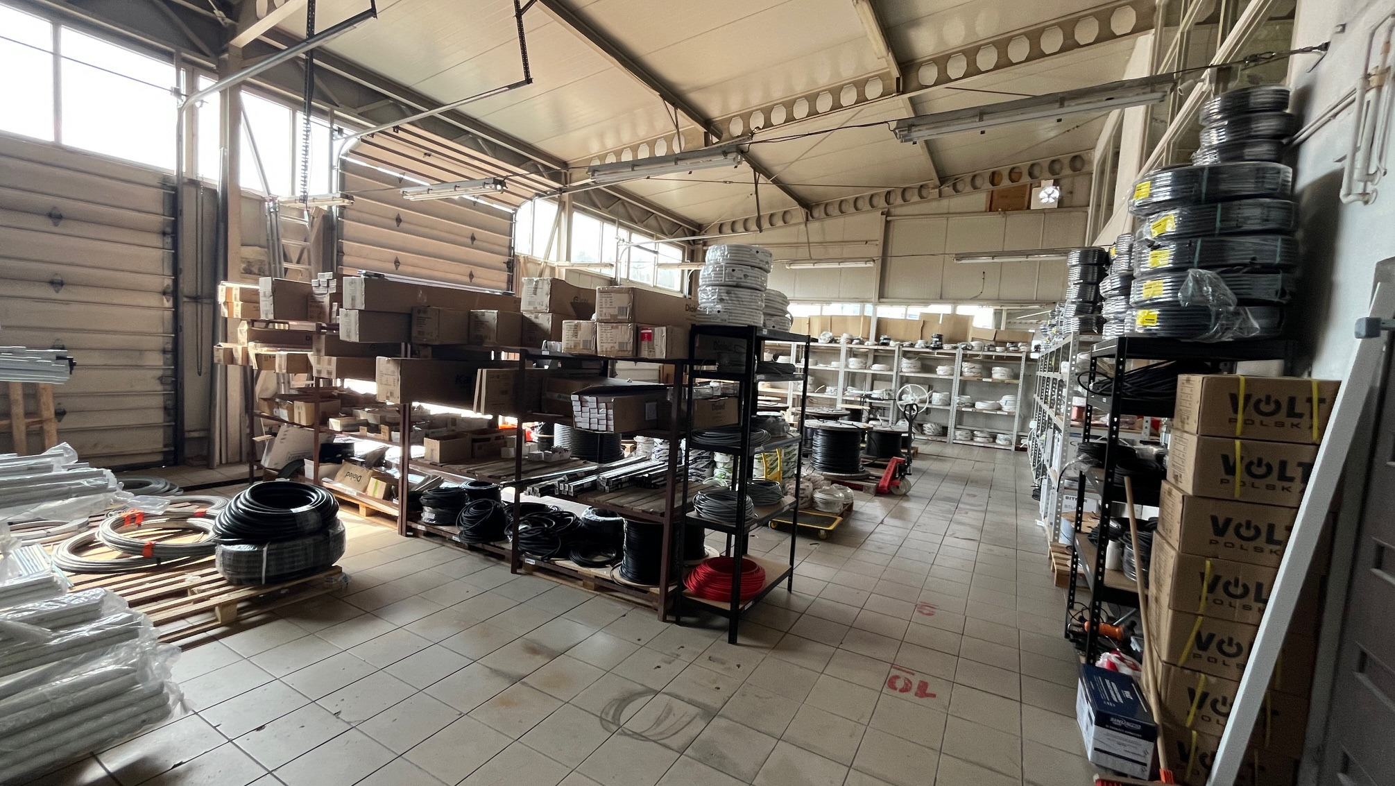 Wnętrze magazynu z półkami pełnymi przewodów i materiałów instalacyjnych, widoczne kartony Volt Polska, oświetlone naturalnym światłem z okien.