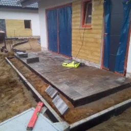 Układanie kostki brukowej na tarasie przy domu z żółtą elewacją, widoczny poziomica, krawężniki i mieszalnik do betonu w tle.