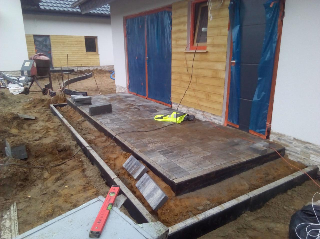 Układanie kostki brukowej na tarasie przy domu z żółtą elewacją, widoczny poziomica, krawężniki i mieszalnik do betonu w tle.