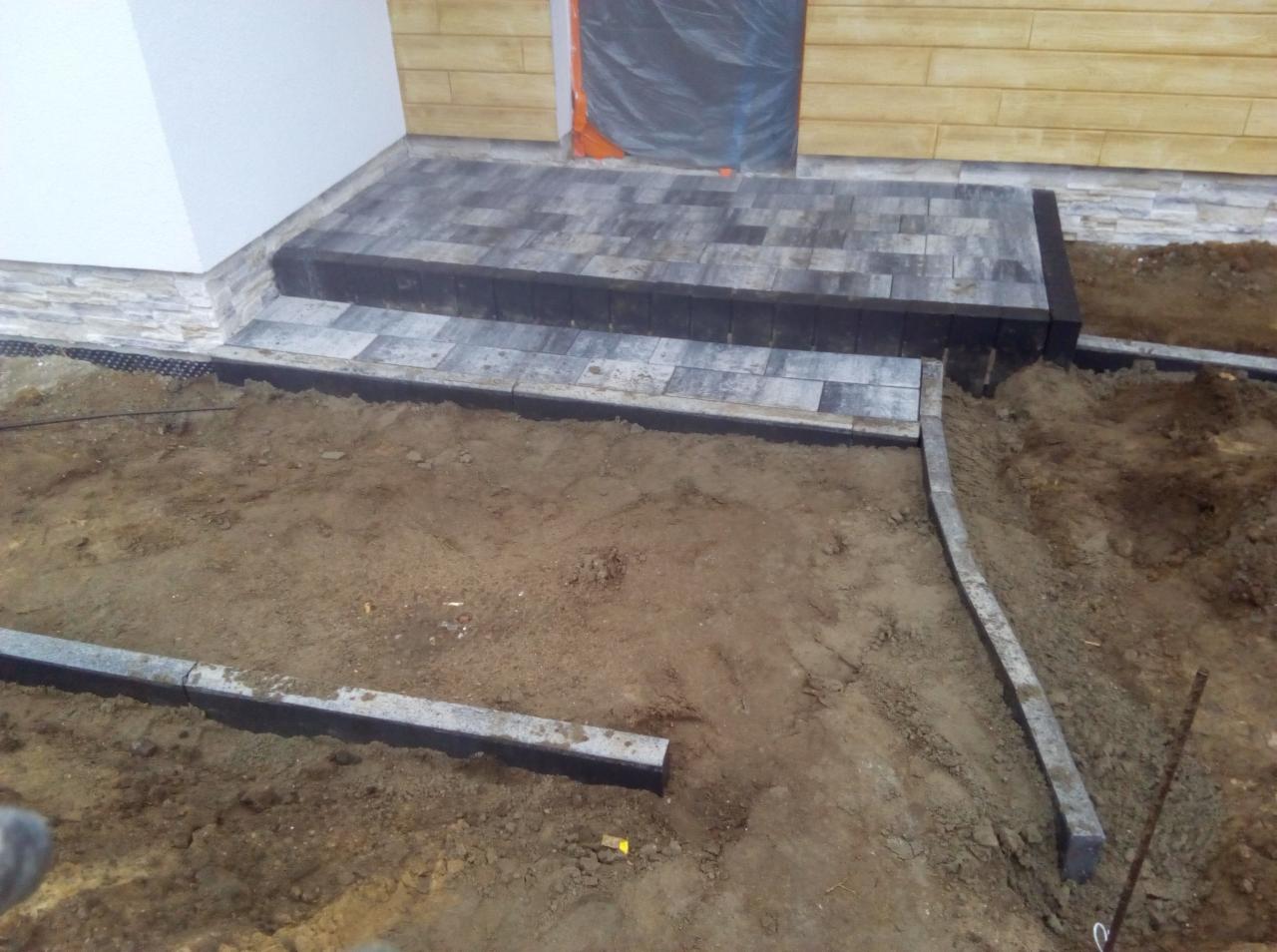 Etap prac brukarskich: ułożona kostka brukowa przed wejściem do domu, widoczne obrzeża i teren przygotowany pod dalsze prace.