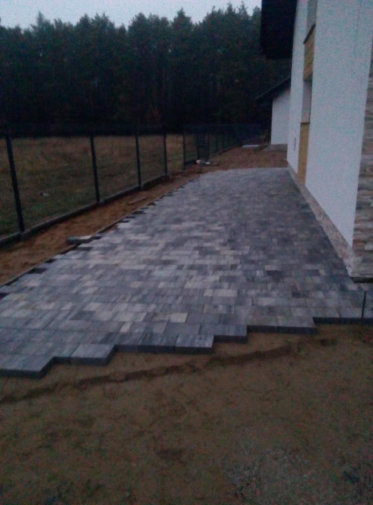 Niedokończona ścieżka z szarej kostki brukowej przy białym budynku, widoczne nierówne krawędzie i przygotowane podłoże z piasku.