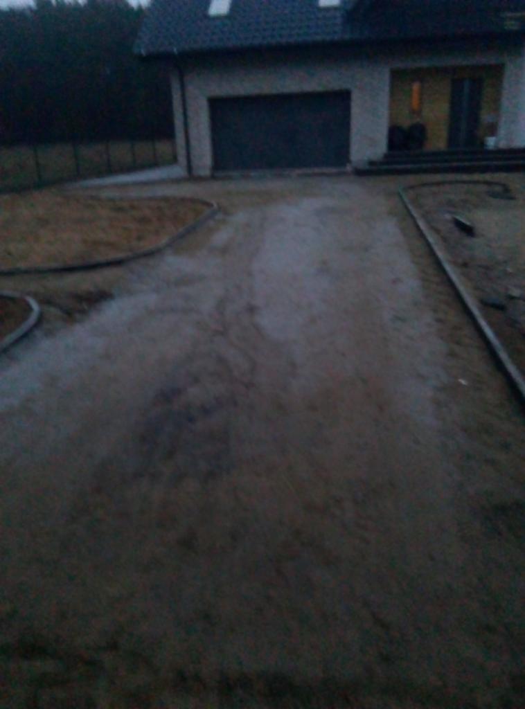 Przygotowanie podłoża pod nawierzchnię przed domem jednorodzinnym, widoczne obrzeża i piasek, w tle budynek z garażem.