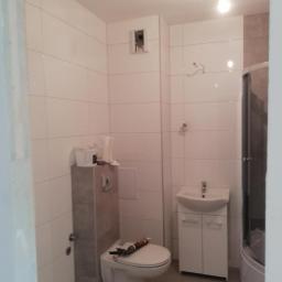 F.H.U Zając Robert - Łazienka w trakcie remontu: białe płytki ścienne, podwieszana toaleta z szarym stelażem, biała szafka z umywalką, kabina prysznicowa w rogu, widoczne narzędzia i materiały budowlane na toalecie.