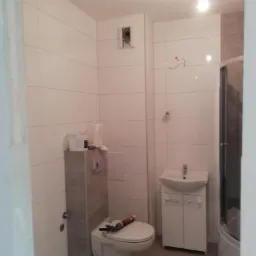 Łazienka w trakcie remontu: białe płytki ścienne, podwieszana toaleta z szarym stelażem, biała szafka z umywalką, kabina prysznicowa w rogu, widoczne narzędzia i materiały budowlane na toalecie.