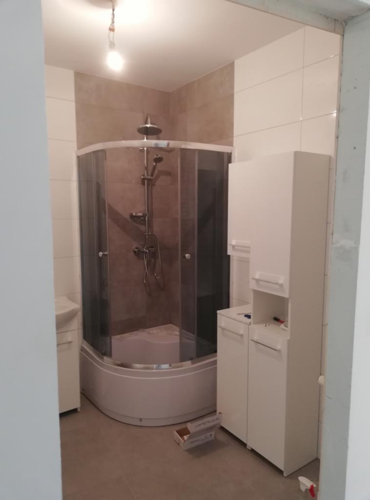 Ukończona łazienka z narożną kabiną prysznicową z brązowymi płytkami i białymi szafkami, widoczne ślady po remoncie na podłodze.