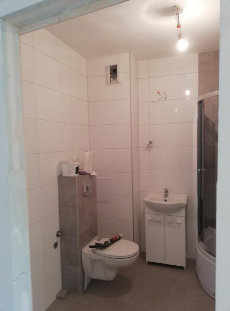 Łazienka w trakcie remontu: białe płytki ścienne, podwieszana toaleta z szarym stelażem, biała szafka z umywalką, kabina prysznicowa w rogu, widoczne narzędzia i materiały budowlane na toalecie.