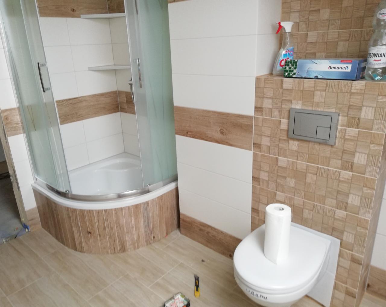 Nowoczesna łazienka po remoncie z kabiną prysznicową z zaokrąglonymi drzwiami, toaletą z rolką papieru i mozaiką na ścianach w odcieniach beżu i brązu.