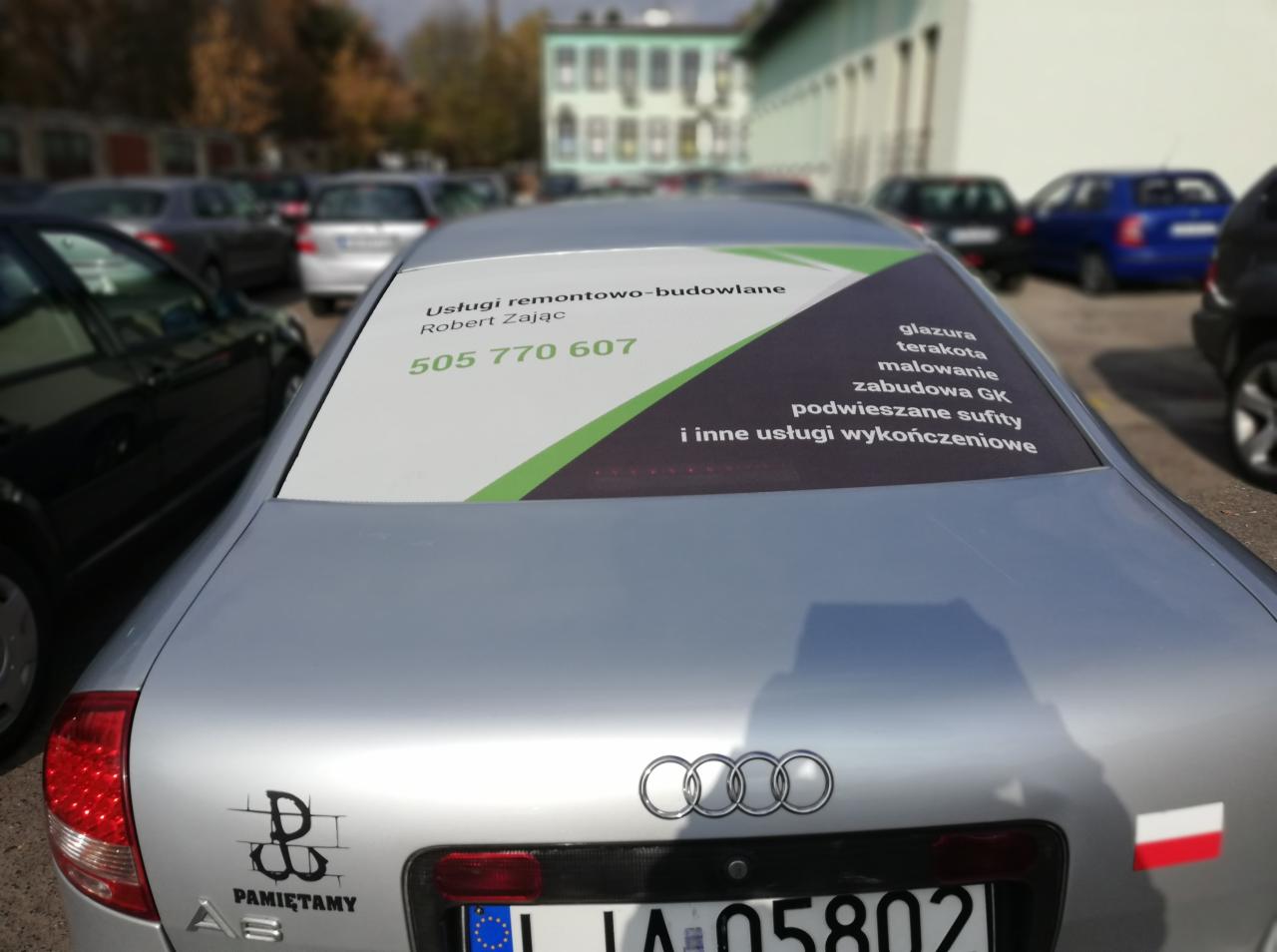 Srebrny samochód Audi A3 z naklejką reklamową usług remontowo-budowlanych na tylnej szybie, zawierającą numer telefonu i listę oferowanych prac wykończeniowych.