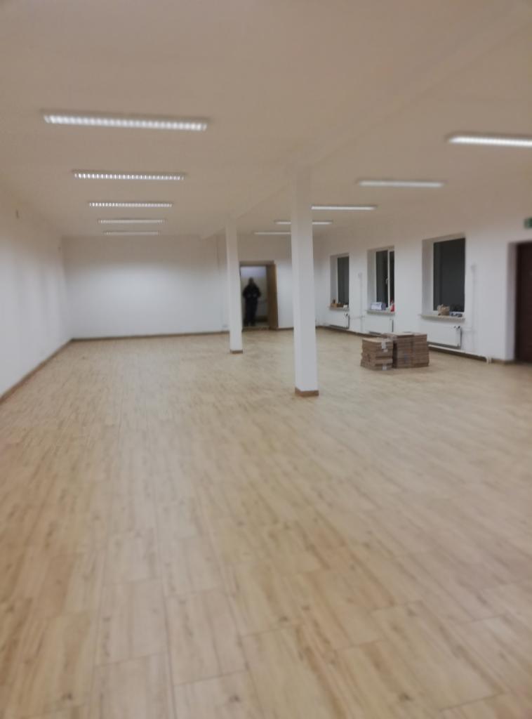 Puste pomieszczenie po remoncie z nową podłogą z płytek imitujących drewno, białe ściany z kolumnami, okna i grzejniki, w tle postać w drzwiach, na podłodze stos płytek.