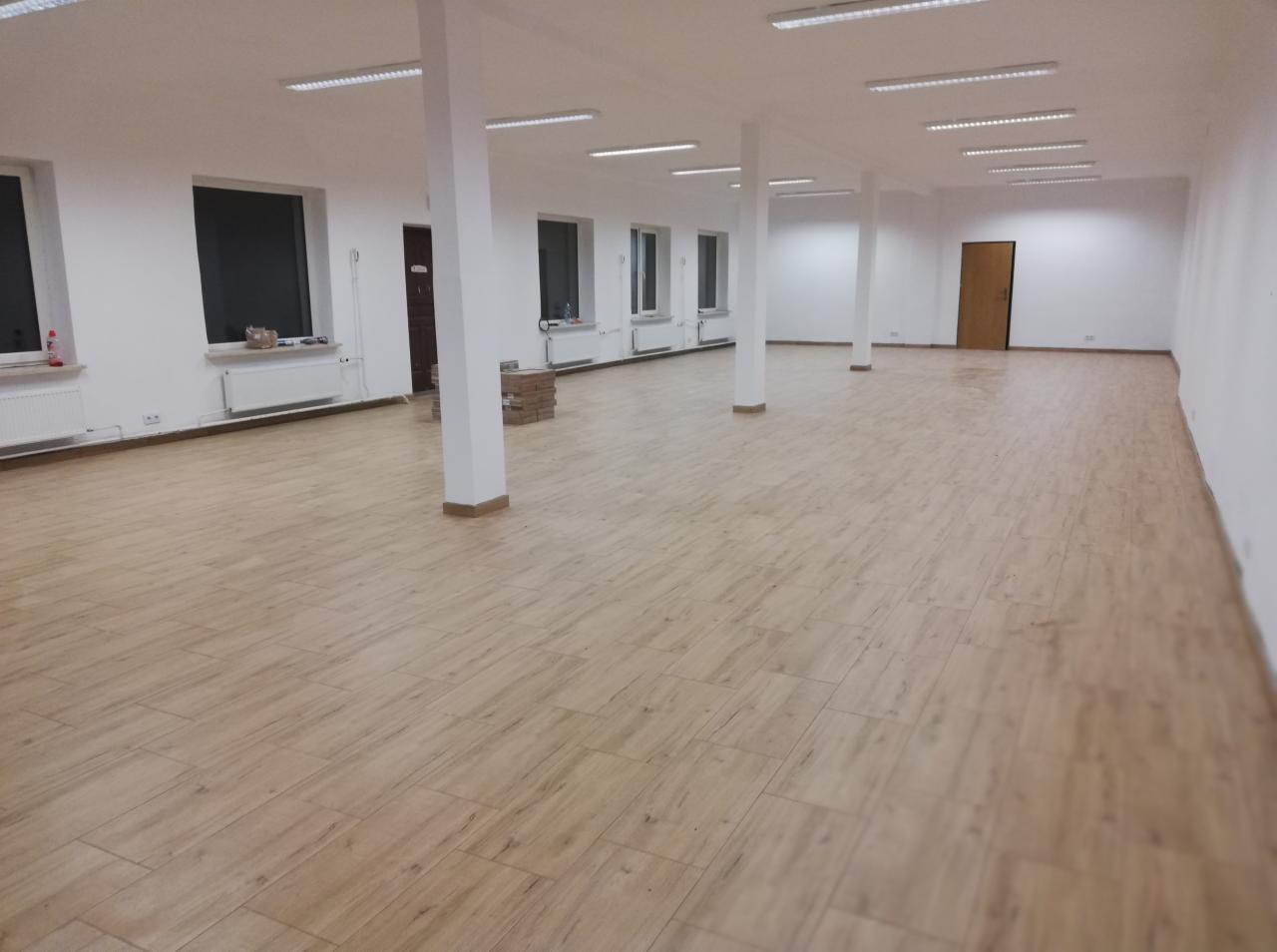 Przestronne wnętrze po remoncie z nową podłogą imitującą drewno ułożoną w jodełkę, białe ściany, kolumny i grzejniki, widoczne okna i drzwi oraz lampy sufitowe.