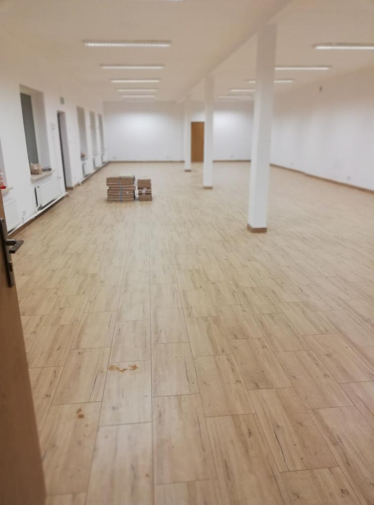 Jasna podłoga z paneli imitujących drewno w przestronnym, pustym pomieszczeniu z białymi ścianami i kolumnami; w tle ułożone kartony z panelami.