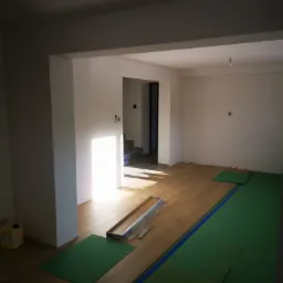 Wnętrze w trakcie układania drewnianej podłogi; widoczne panele podłogowe, zielona folia ochronna, listwy przypodłogowe w opakowaniu i pojemnik z klejem