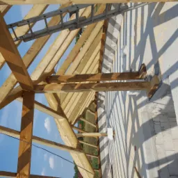 Drewniana konstrukcja więźby dachowej w trakcie budowy, widoczna drabina oparta o konstrukcję, ściany z białego betonu komórkowego, niebieskie niebo w tle.