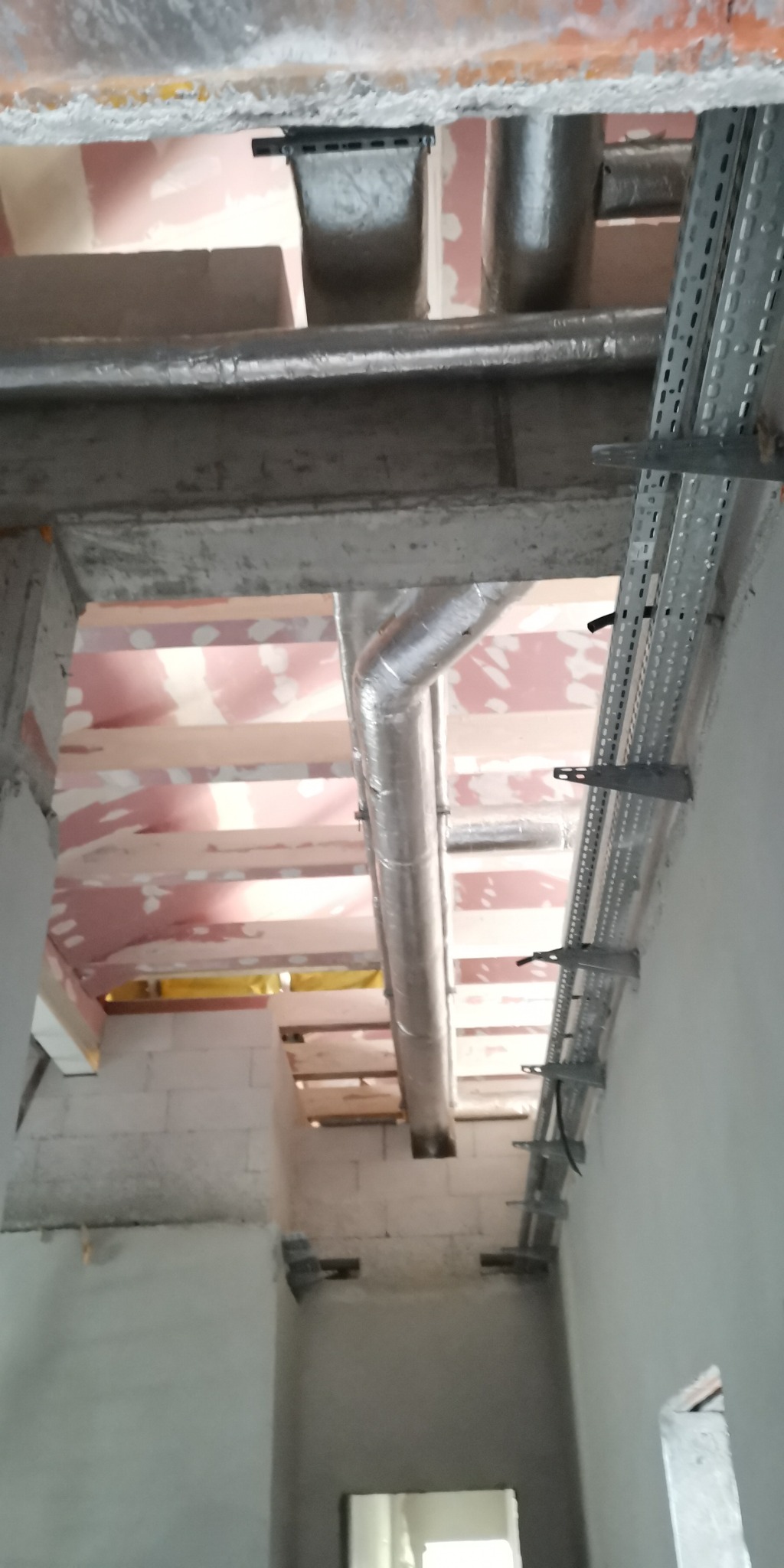 Instalacja wentylacyjna z rurami aluminiowymi i metalowe korytka kablowe zamocowane do sufitu w surowym stanie budynku, widoczne białe bloczki i izolacja stropu.