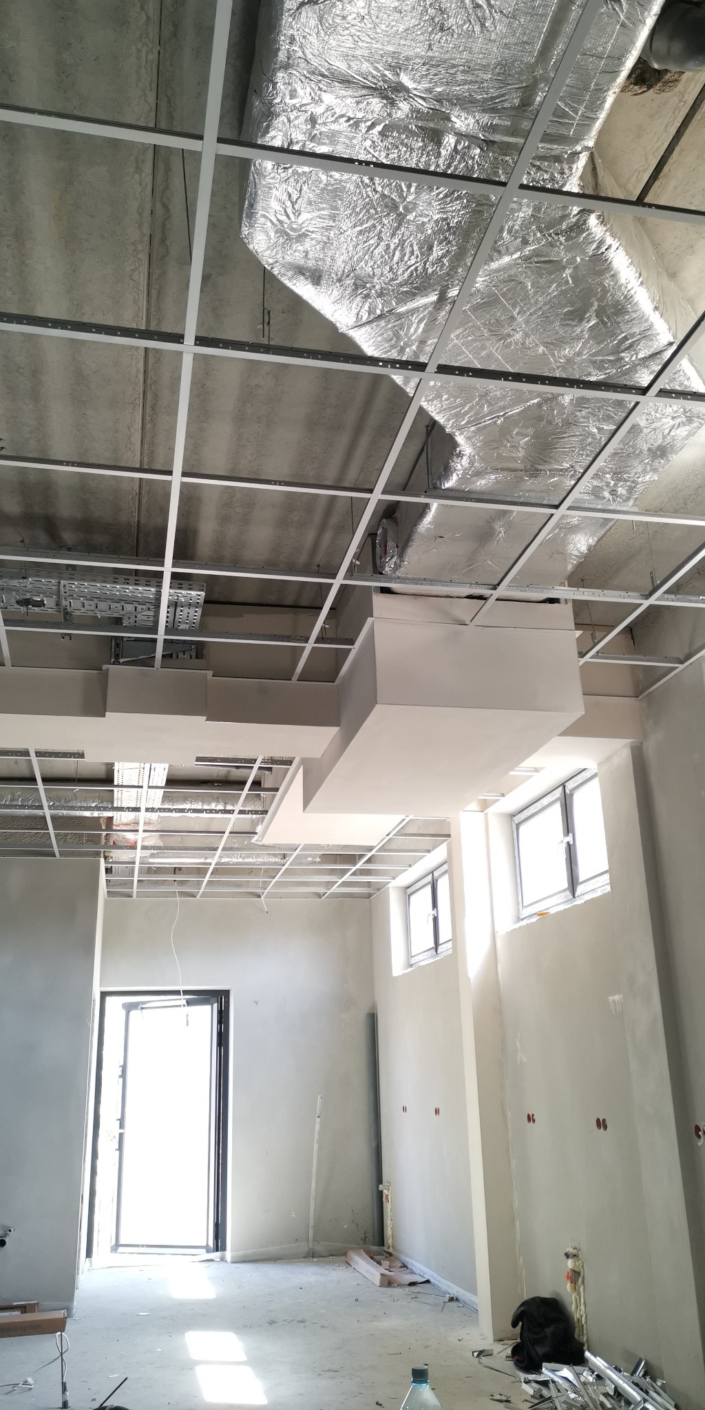 Montaż sufitu podwieszanego z płyt kartonowo-gipsowych, widoczne profile, izolacja i elementy instalacji wentylacyjnej w surowym pomieszczeniu z oknami.