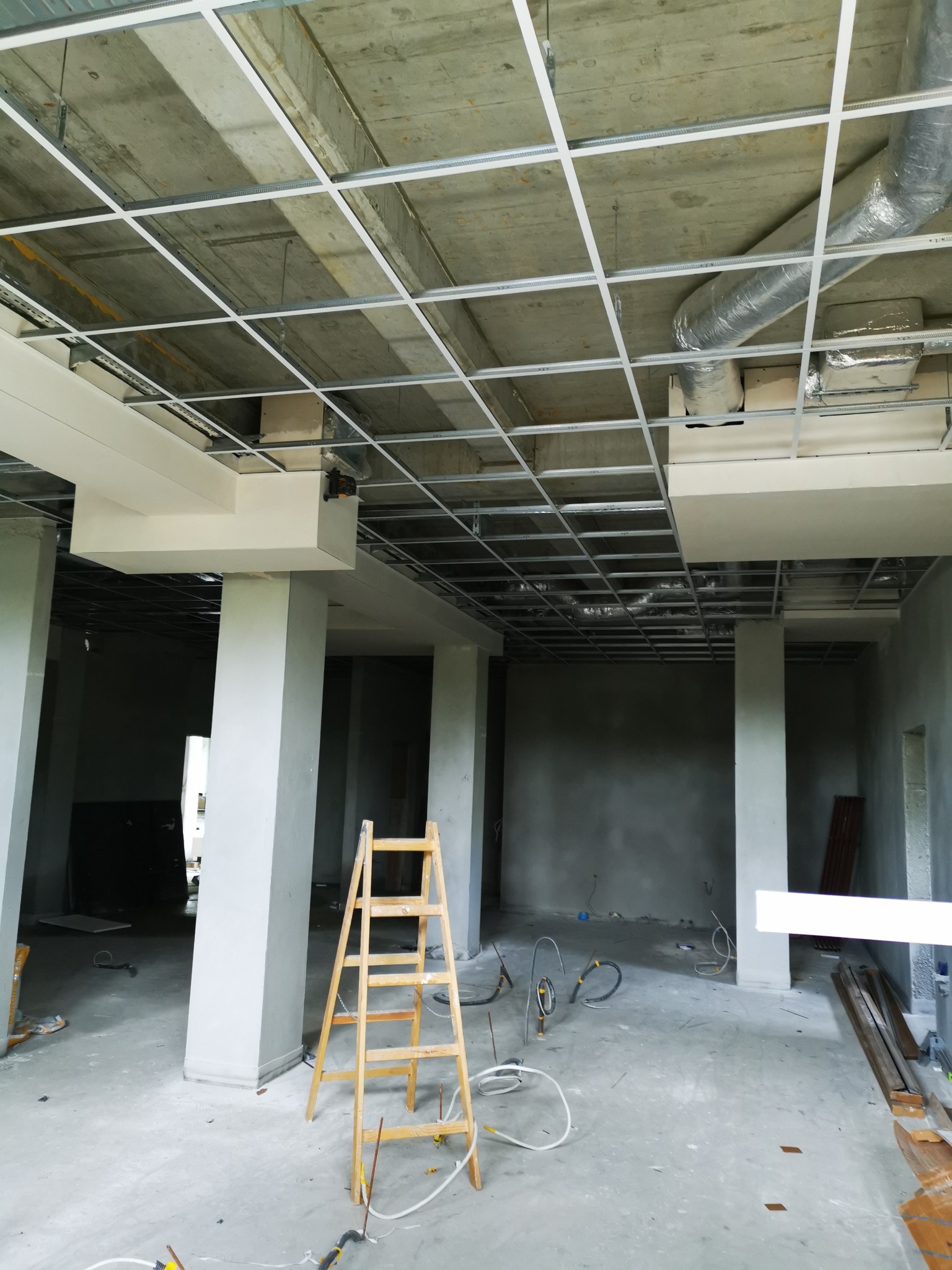 Wnętrze w trakcie prac wykończeniowych: widoczna metalowa konstrukcja sufitu podwieszanego, częściowo zamontowane białe elementy sufitu, drewniana drabina i kable na podłodze, surowy betonowy strop.