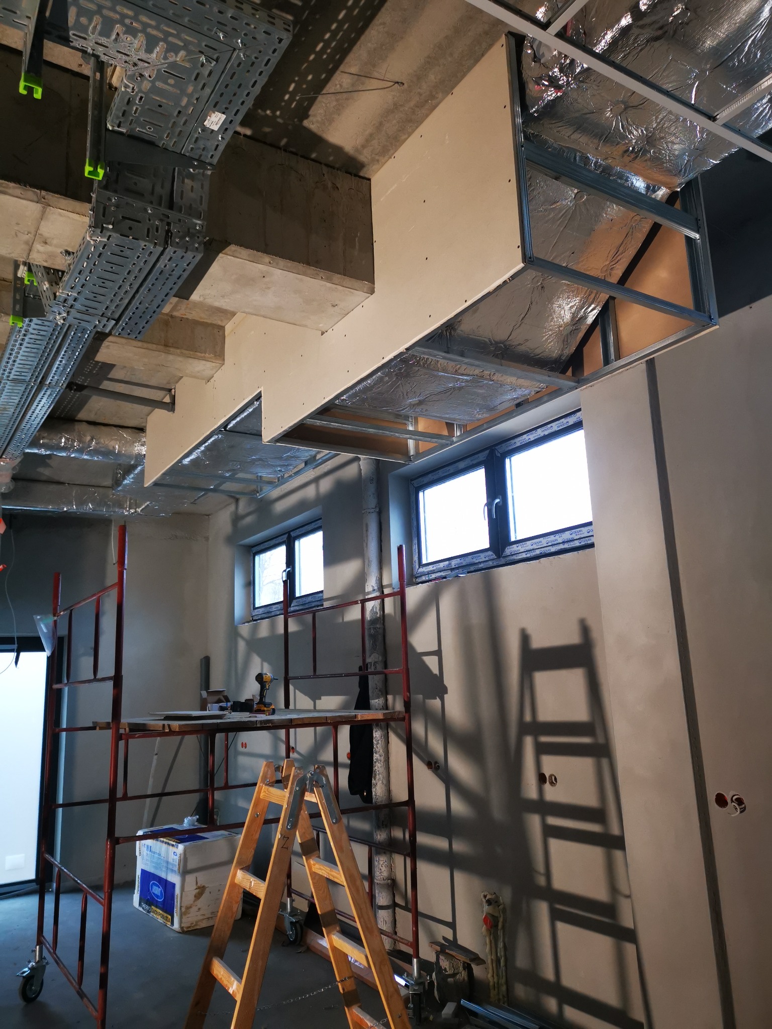 Montaż sufitu podwieszanego z płyt gipsowo-kartonowych w pomieszczeniu z oknami, widoczne profile metalowe, izolacja, rusztowanie i drabina.
