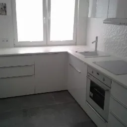 Jasna kuchnia z białymi frontami szafek, szarą podłogą i oknem z widokiem na zewnątrz, z płytkami dekoracyjnymi na ścianie.