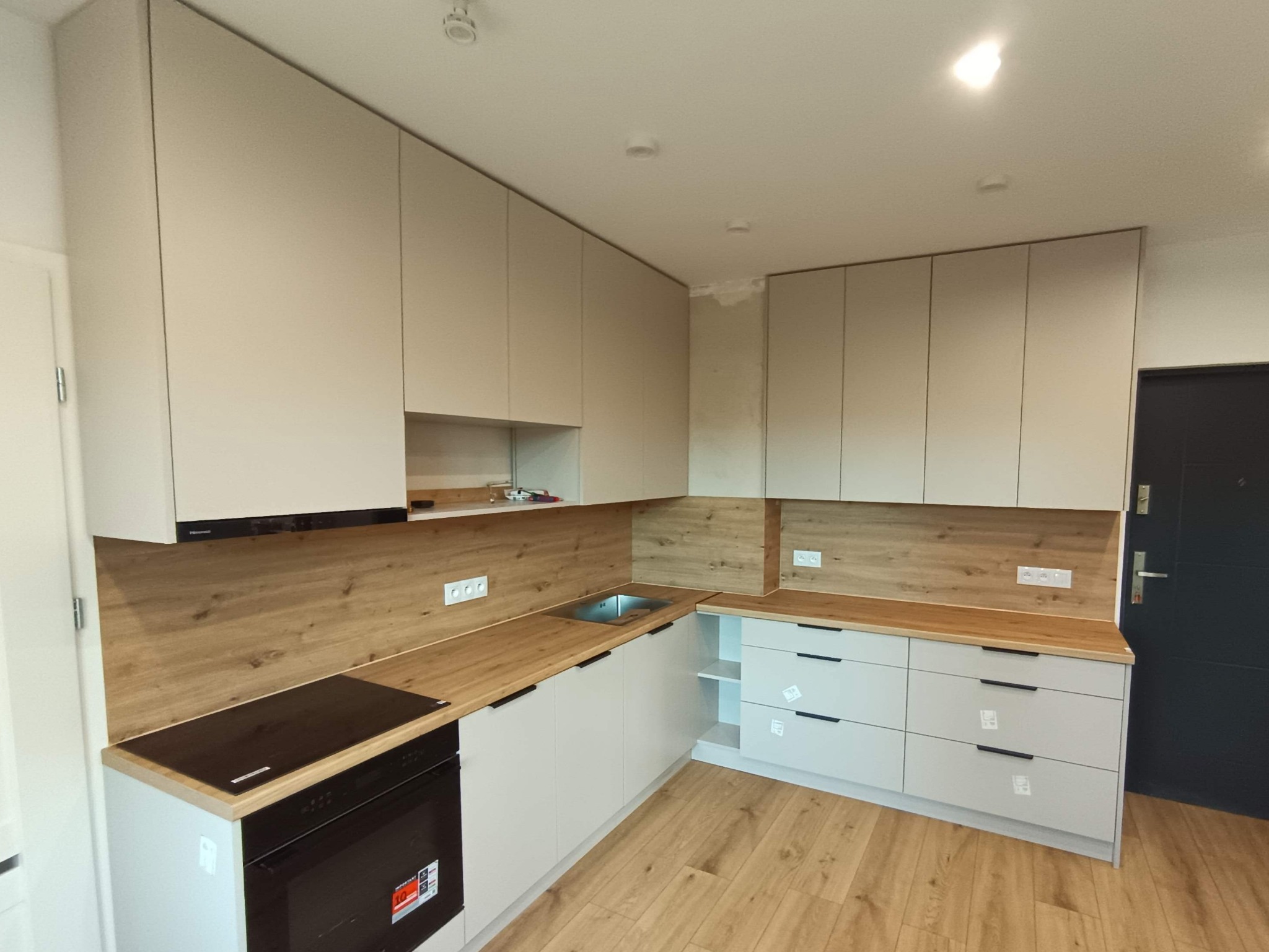 Jasna kuchnia z drewnianym blatem i panelami, minimalistyczne uchwyty, nowoczesny design, widok z perspektywy wnętrza, oświetlenie sufitowe.