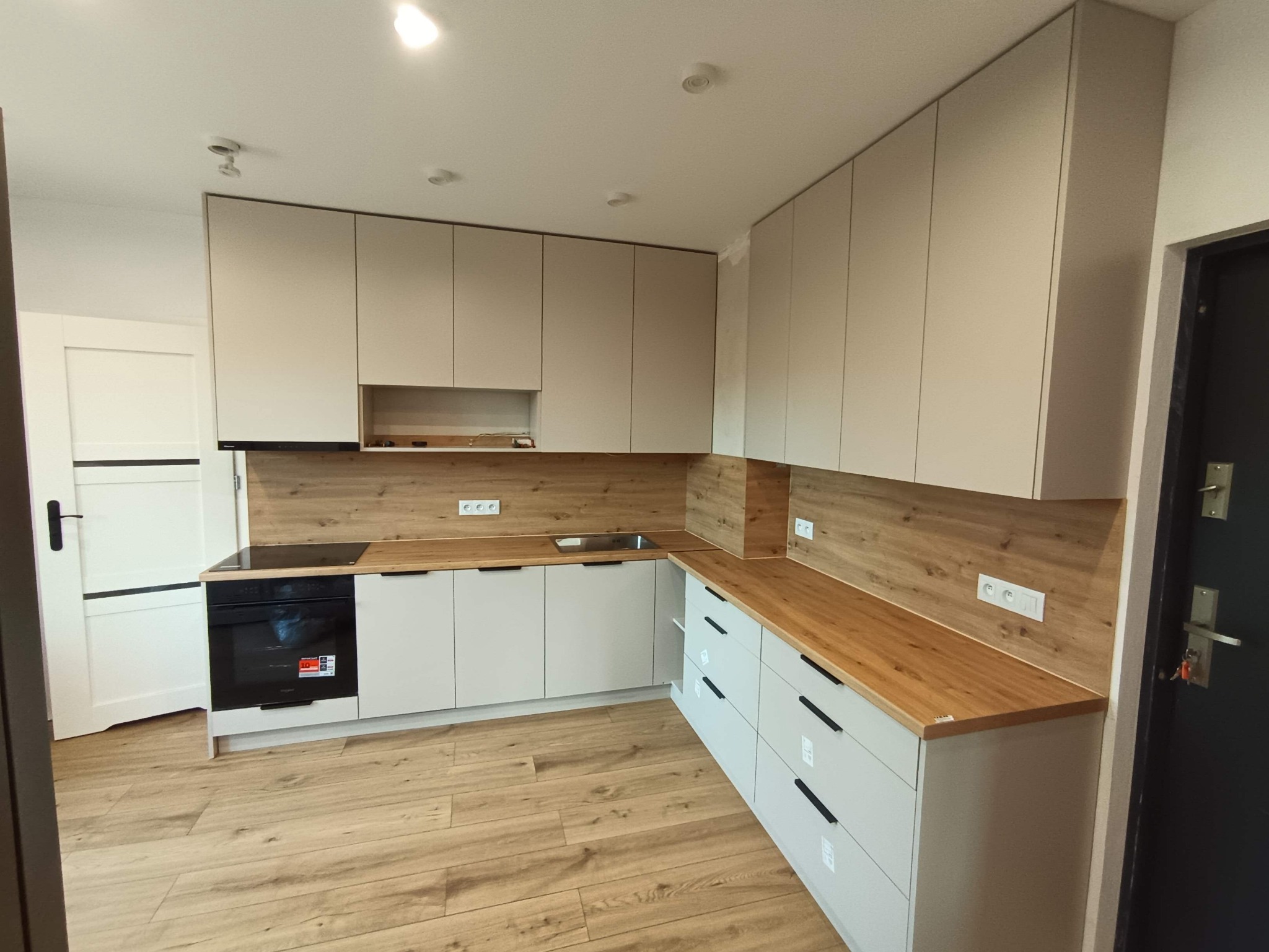 Jasna kuchnia z drewnianym blatem i panelami. Minimalistyczny design, fronty bez uchwytów. Widoczna płyta indukcyjna i piekarnik. Podłoga drewniana.