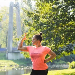 Kobieta w sportowym stroju, pozująca na tle parku z mostem w oddali, wykonująca element ćwiczeń rozciągających.