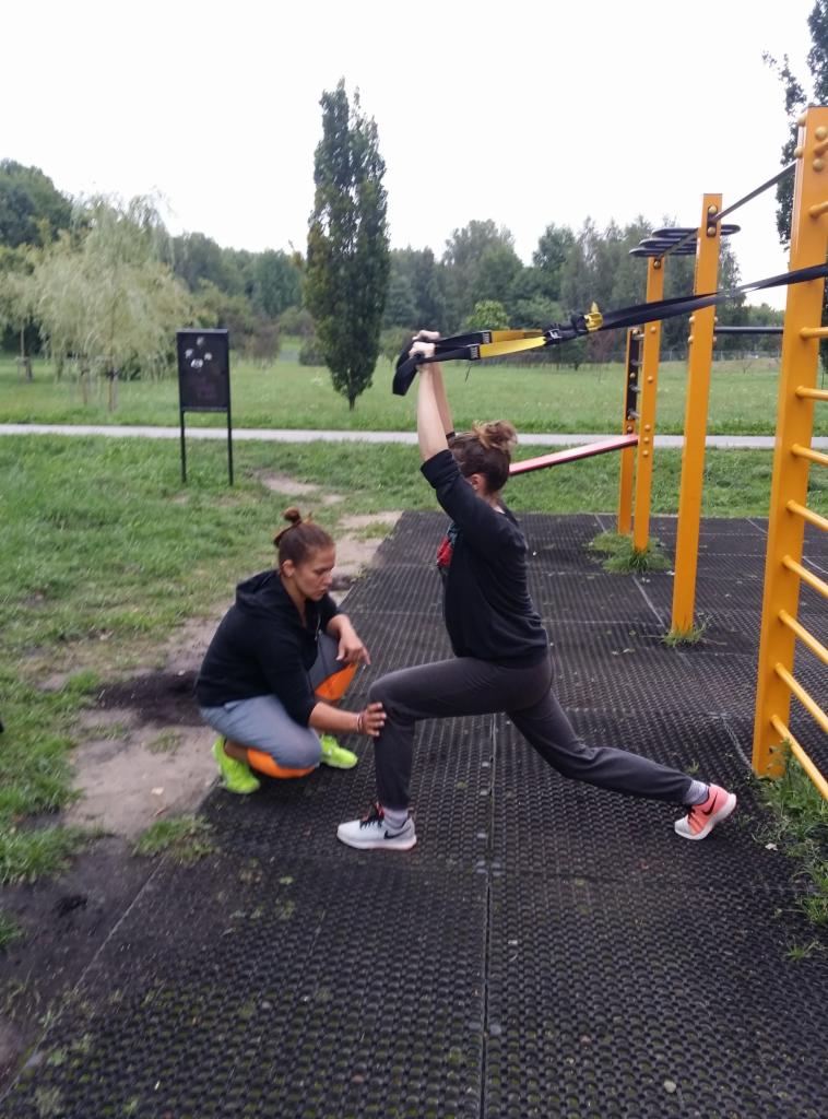 Kobieta wykonuje ćwiczenie w parku na drabinkach, pod nadzorem drugiej kobiety w jaskrawozielonych butach.