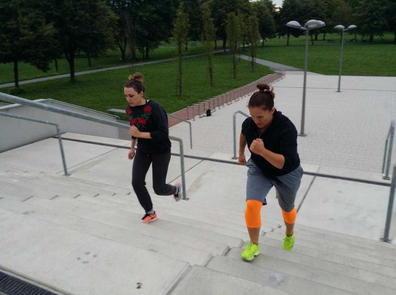Dwie kobiety w sportowych strojach wbiegają po betonowych schodach w parku miejskim, oświetlonych przez wysokie lampy.