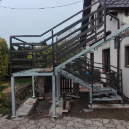 Konstrukcja schodów zewnętrznych ze stali czarnej w trakcie montażu przy budynku mieszkalnym, widoczne elementy mocujące i surowy stan otoczenia.