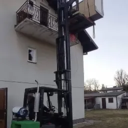 Wózek widłowy Clark podnosi meble na balkon domu jednorodzinnego, widoczny operator w kabinie i butla gazowa na masce.