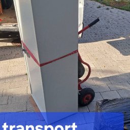 M. MARIO TRANS MARIUSZ ZIEŃ - Transport sejfu na wózku transportowym, zabezpieczonego czerwoną taśmą, przygotowanego do załadunku do busa. Widoczne rękawice robocze na sejfie.