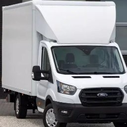 M. MARIO TRANS MARIUSZ ZIEŃ - Biały Ford Transit z zabudową kontenerową na tle ceglanej ściany i okien. Widok z przodu, lekko z boku. Czysty, zadbany pojazd.