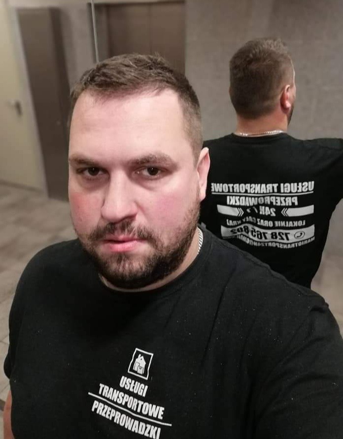 Mężczyzna w czarnej koszulce z logo firmy transportowej i przeprowadzkowej, z drugim mężczyzną w tle, obaj stoją w pobliżu windy.