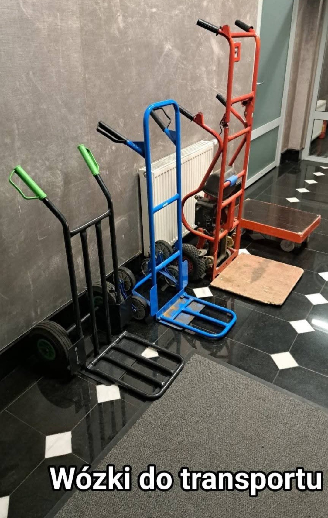 Zestaw wózków transportowych: czarny z zielonymi uchwytami, niebieski z sześcioma kółkami oraz czerwony schodołaz z silnikiem spalinowym, ustawione na czarno-białej posadzce w korytarzu.
