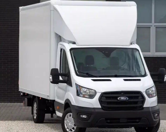 Biały Ford Transit z zabudową kontenerową na tle ceglanej ściany i okien. Widok z przodu, lekko z boku. Czysty, zadbany pojazd.