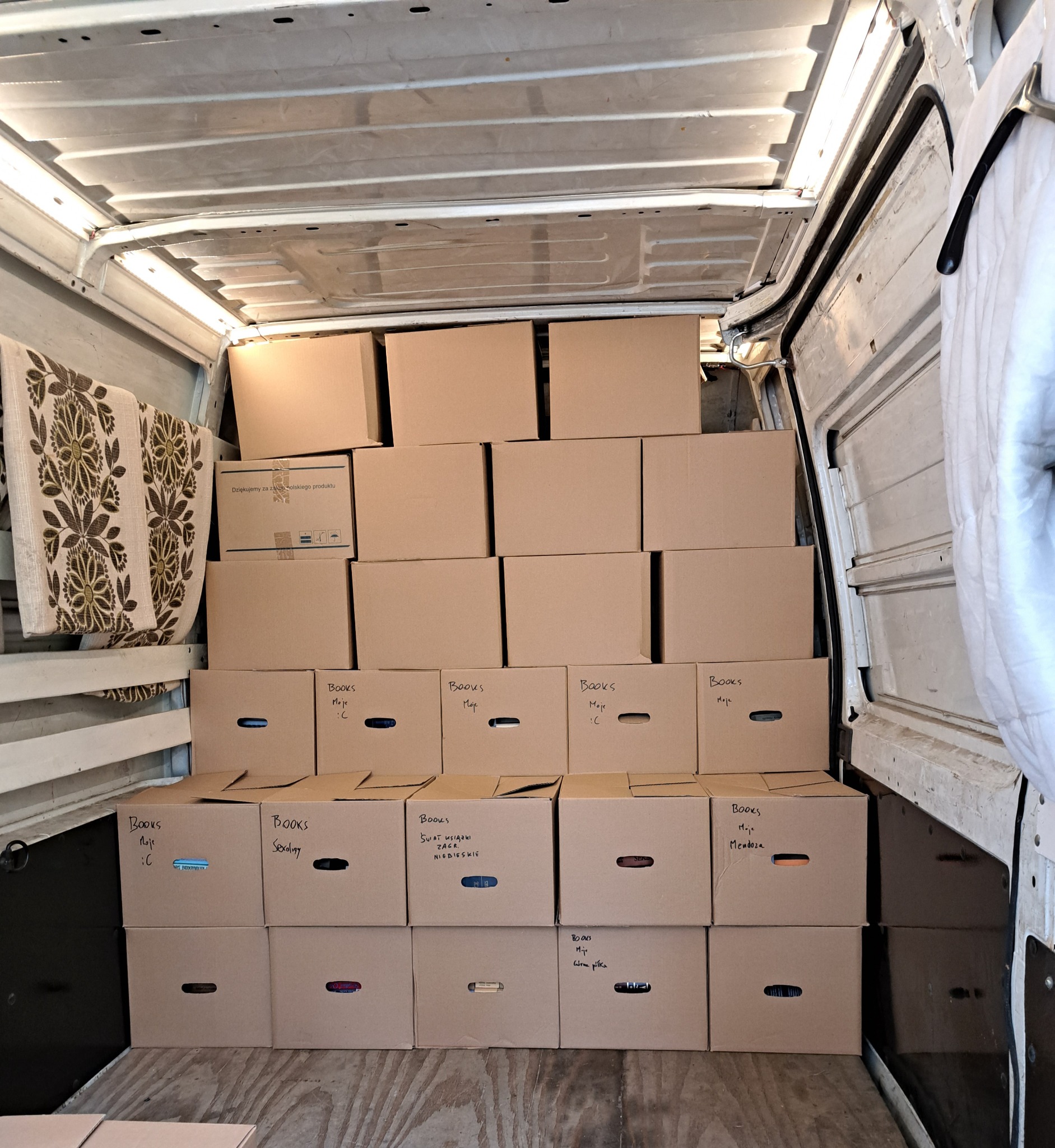 Wnętrze busa wypełnione kartonami z napisem 'Books' oraz nazwami własnymi, ułożonymi w piramidę, widoczny fragment wzorzystej tkaniny po lewej stronie.