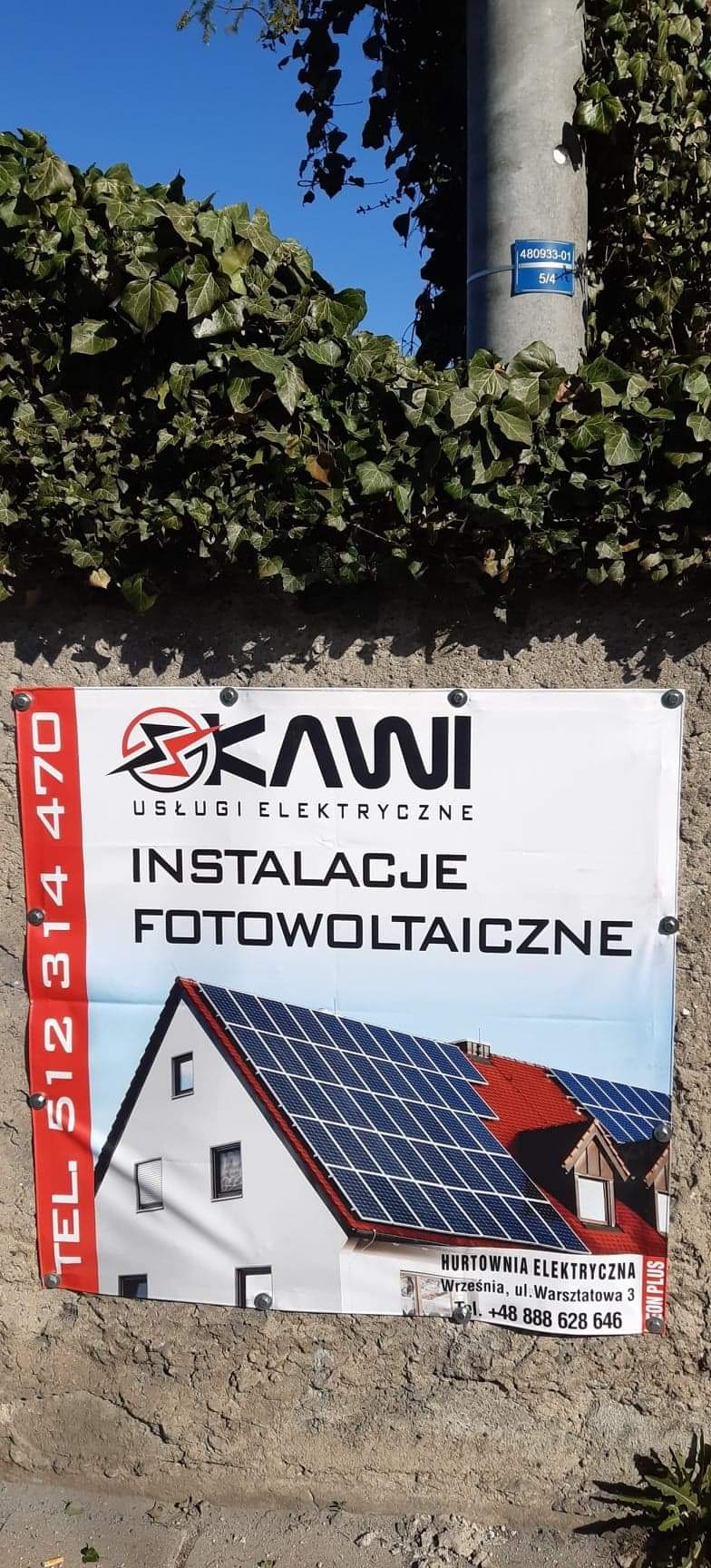 Billboard firmy Skawi oferującej usługi elektryczne i instalacje fotowoltaiczne, z ilustracją domu z panelami słonecznymi na dachu, umieszczony na betonowym murze obok słupa z numerem.