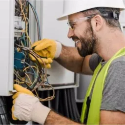 Uśmiechnięty mężczyzna w kasku i okularach ochronnych, w żółtej kamizelce i rękawicach, pracuje przy skrzynce elektrycznej z licznymi kablami.