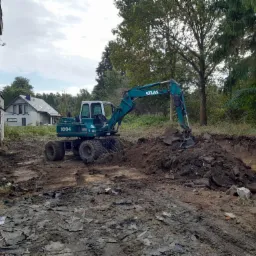 Niebieska koparka Atlas 1004 w trakcie prac ziemnych na terenie budowy, z widocznymi fragmentami gruzu i starego budynku w tle.