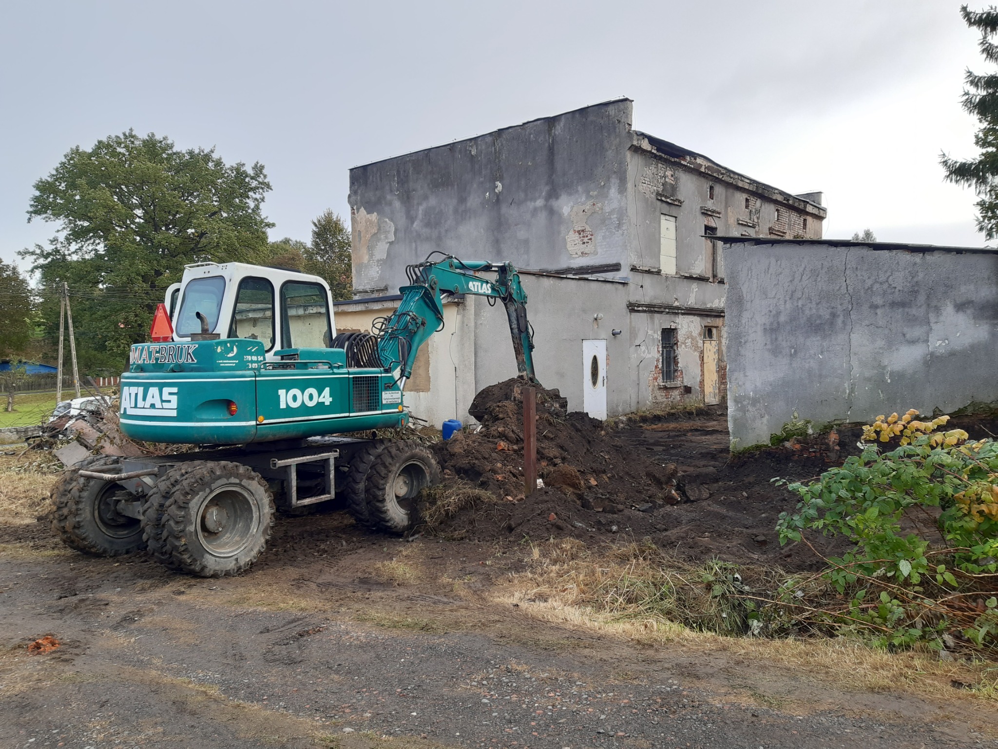 Zielona koparka Atlas 1004 na tle częściowo rozebranego, szarego budynku z odpadającym tynkiem, z wykopaną ziemią i sterczącym metalowym słupkiem.