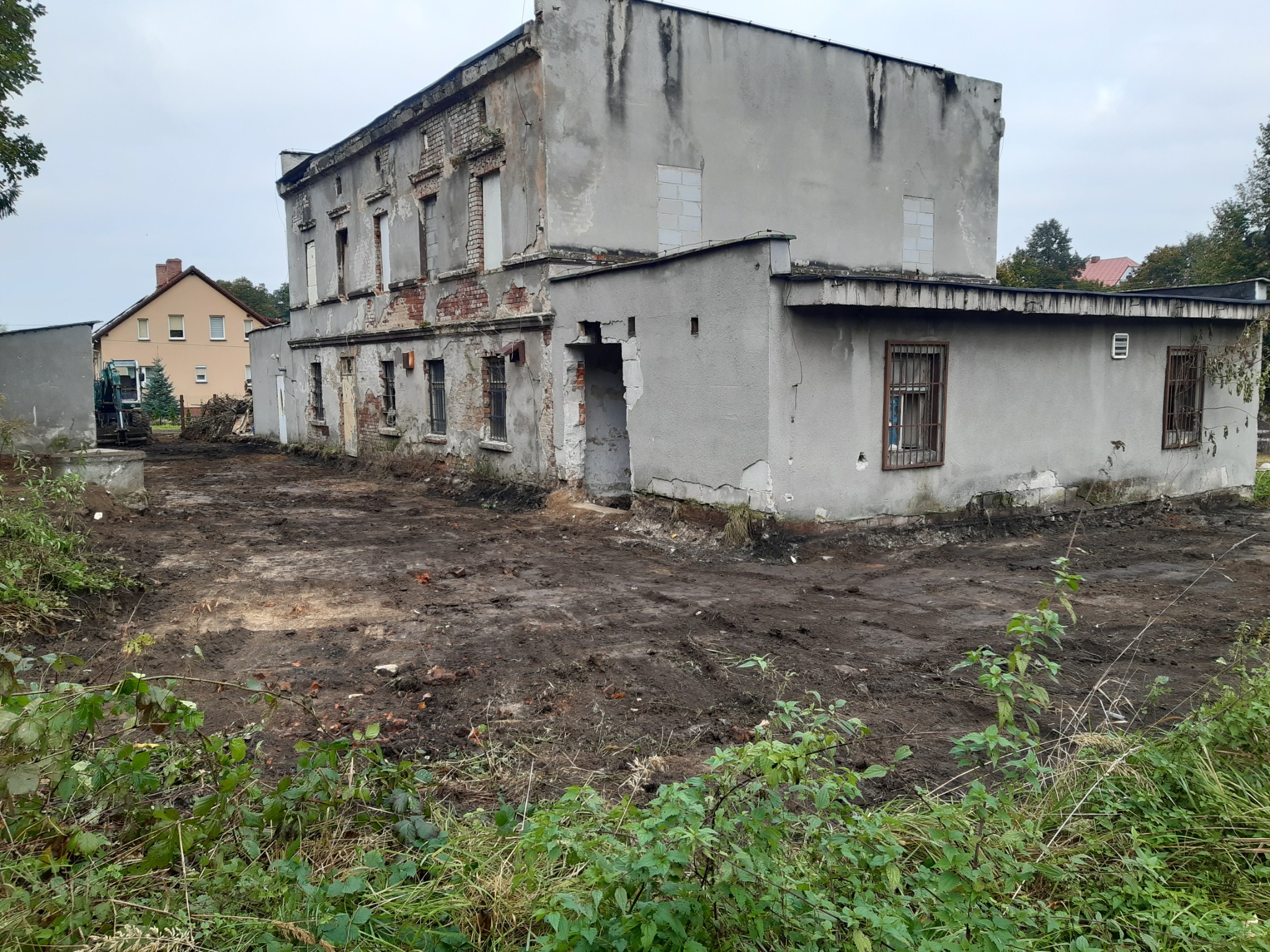 Przygotowany teren po rozbiórce starego, dwukondygnacyjnego budynku z odsłoniętą cegłą i resztkami tynku, z widocznym sprzętem budowlanym w tle.
