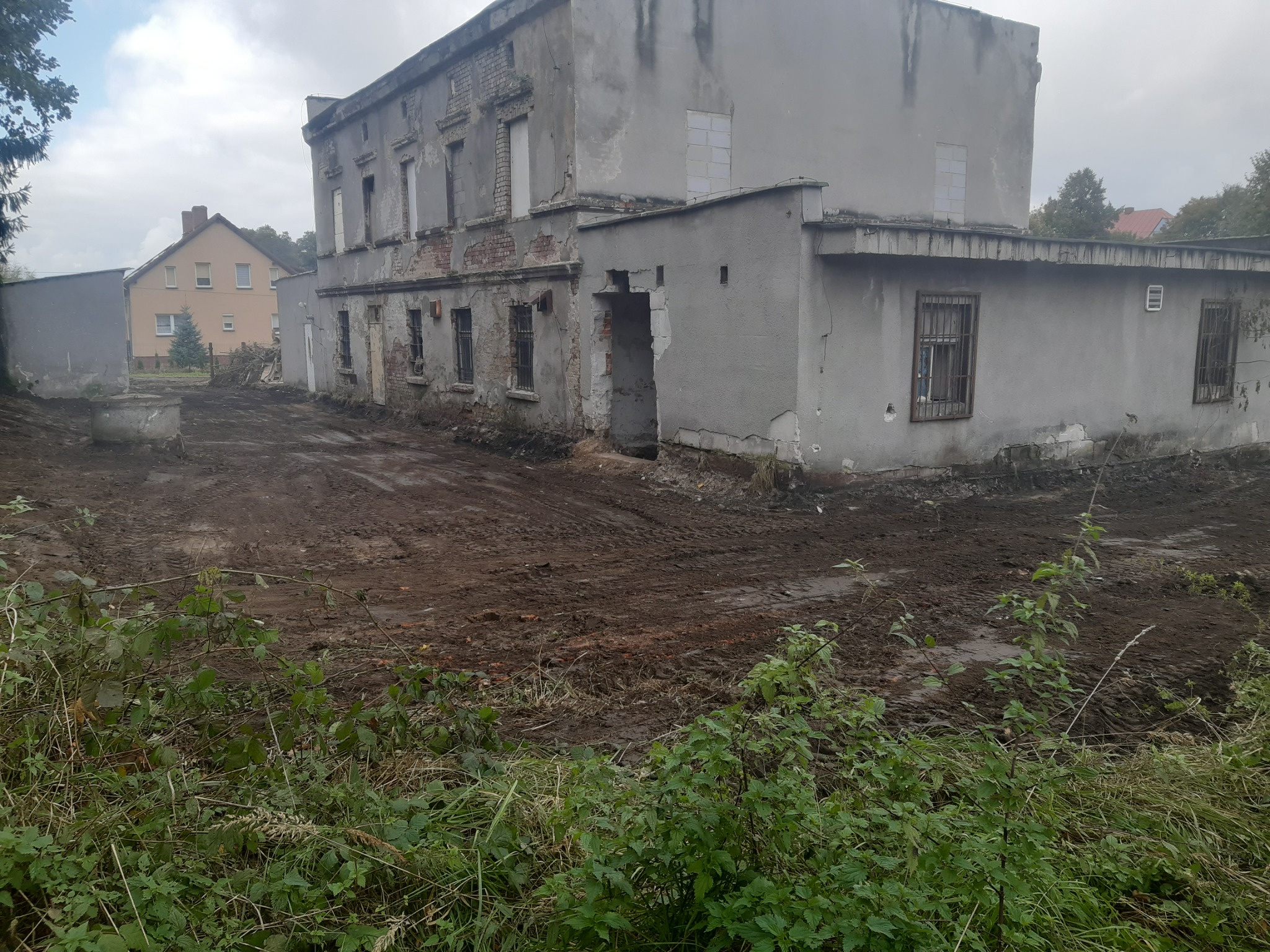 Zdjęcie przedstawia częściowo rozebrany, dwukondygnacyjny budynek z odsłoniętą cegłą i zakratowanymi oknami, otoczony błotnistym terenem z widocznymi śladami opon i zieloną roślinnością...