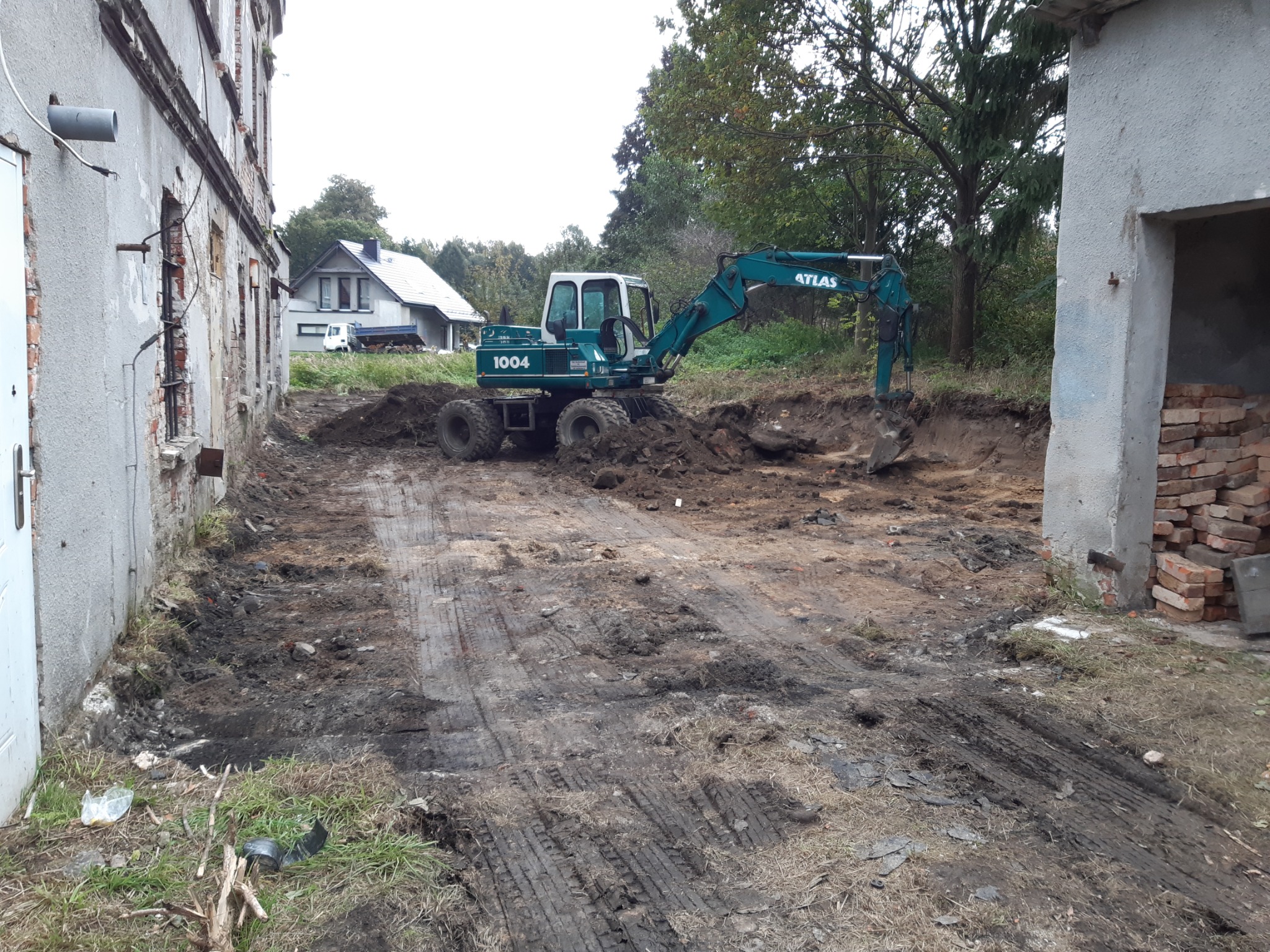 Zielono-biała koparka kołowa Atlas 1004 w trakcie prac ziemnych na placu budowy, widoczne ślady opon w błocie i fragmenty gruzu, w tle budynek z cegły i dom mieszkalny.