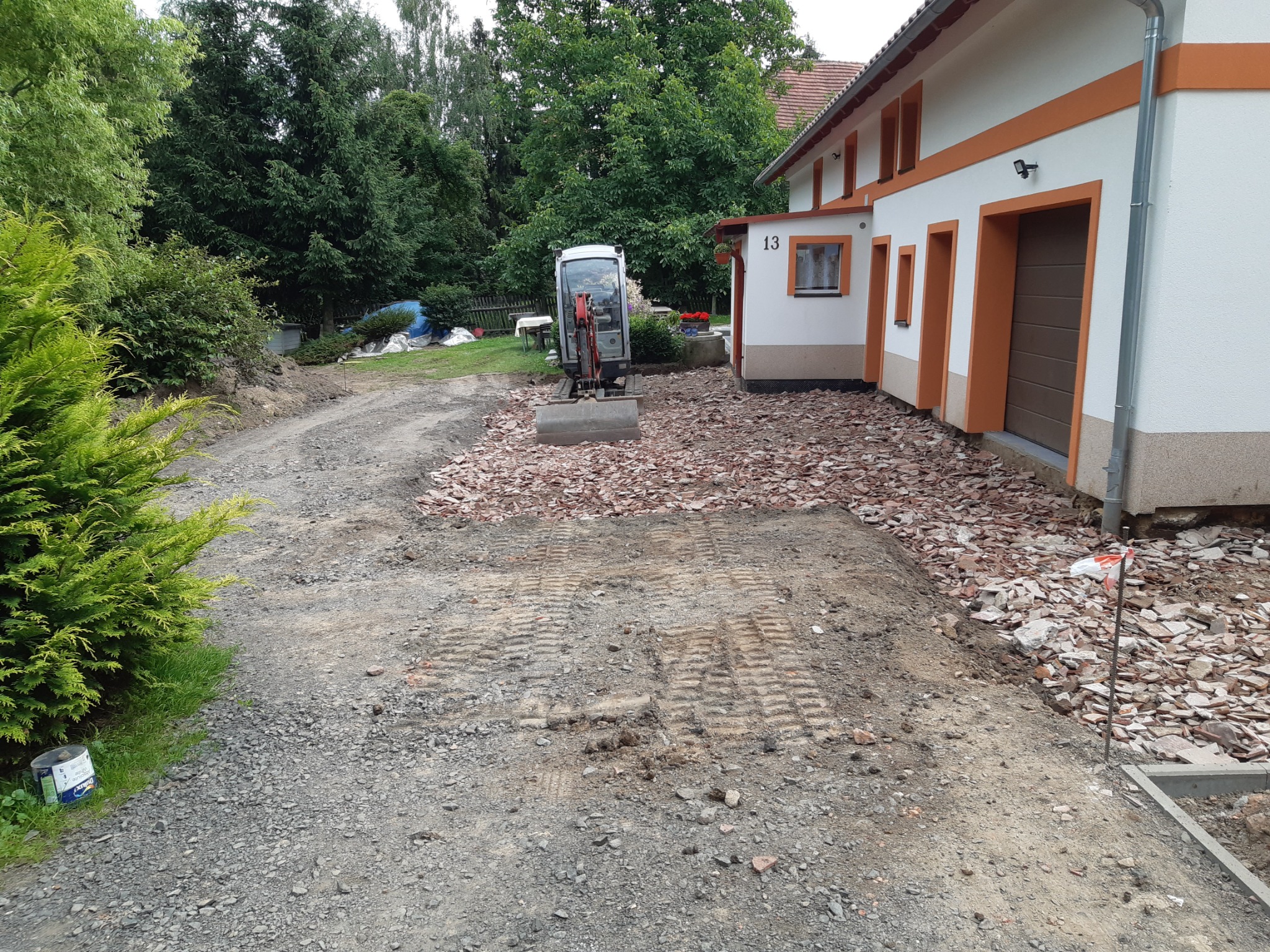 Mini koparka na placu budowy przed domem z numerem 13, teren przygotowany pod wykop z widocznymi śladami gąsienic, obok rozrzucone fragmenty gruzu, w tle zieleń drzew.